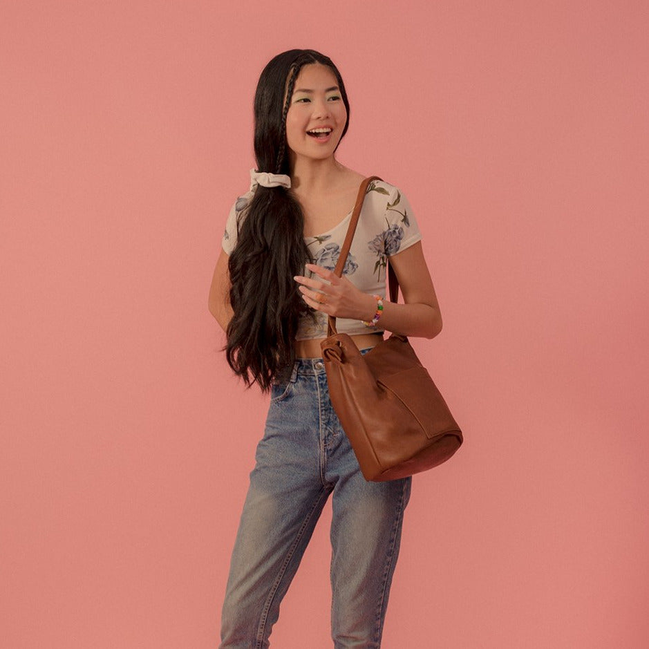 the erin templeton cross body bucket bag in caramel, shown on a model on a pink background. the model is wearing a cream coloured floral top and mid wash levi's jeans