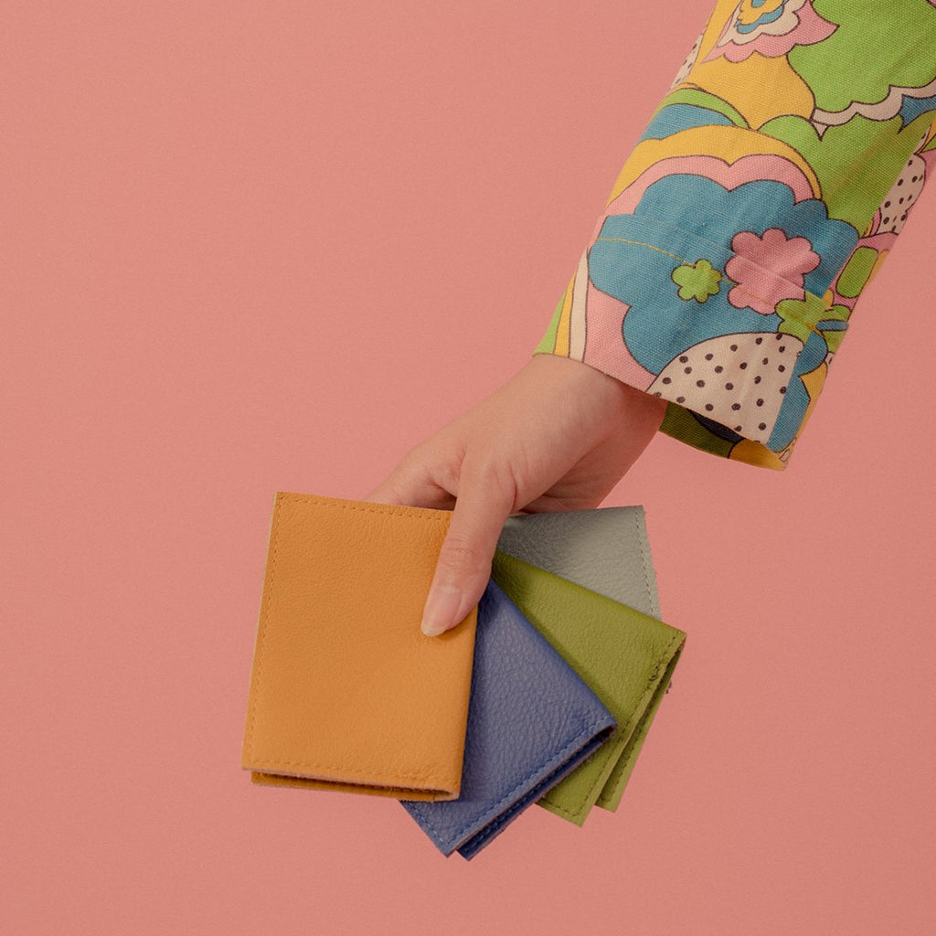 an arm clad in a colourful sleeve holding four folded leather card cases in mustard, lilac, kermit and toothpaste leathers over a pink background
