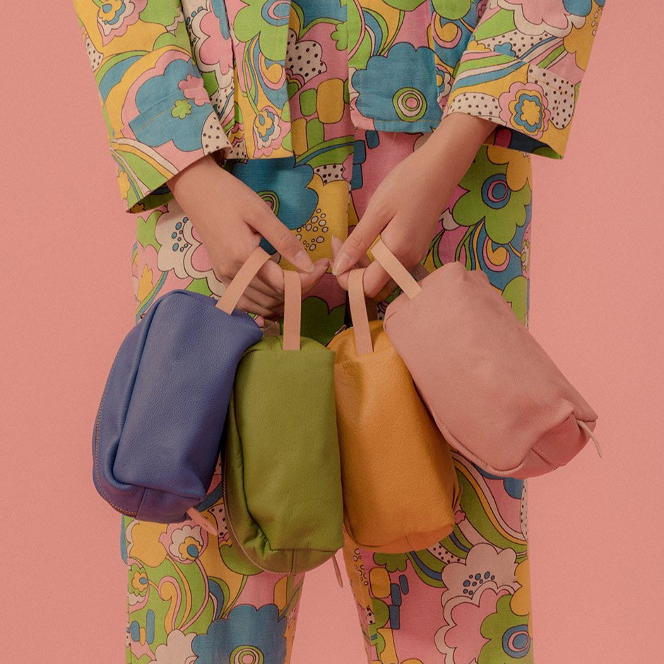 a model wearing a colourfully printed suit holding four leather dopp kits or toiletries bags in blue, kermit, mustard and blush leathers, each with a veg tan leather handle in front of a pink background