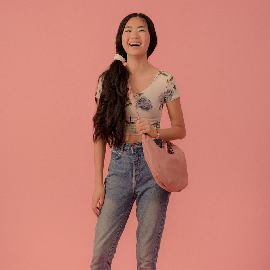 a large hobo leather purse in blush leather with a zip closure, worn cross body on a model wearing a blue and cream floral tee and jeans in front of a  pink back drop