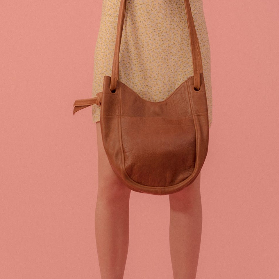 a small leather tote with a large front pocket, double straps and a magnetic closure in caramel leather, held in front of a model wearing a yellow dress in front of a pink backdrop