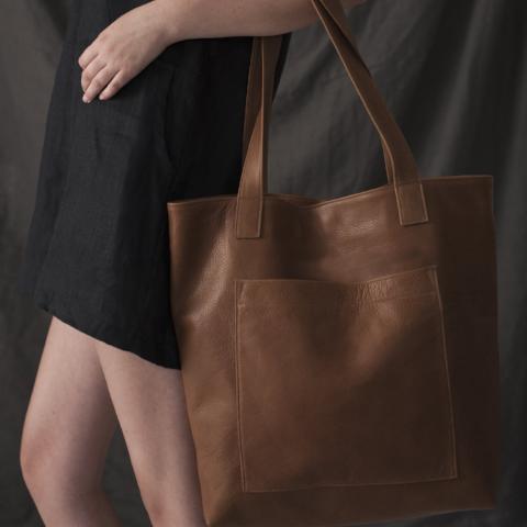a large leather tote with a large centred patch-style pocket shown in caramel leather, worn on the arm of a model in a black dress against a black backdrop