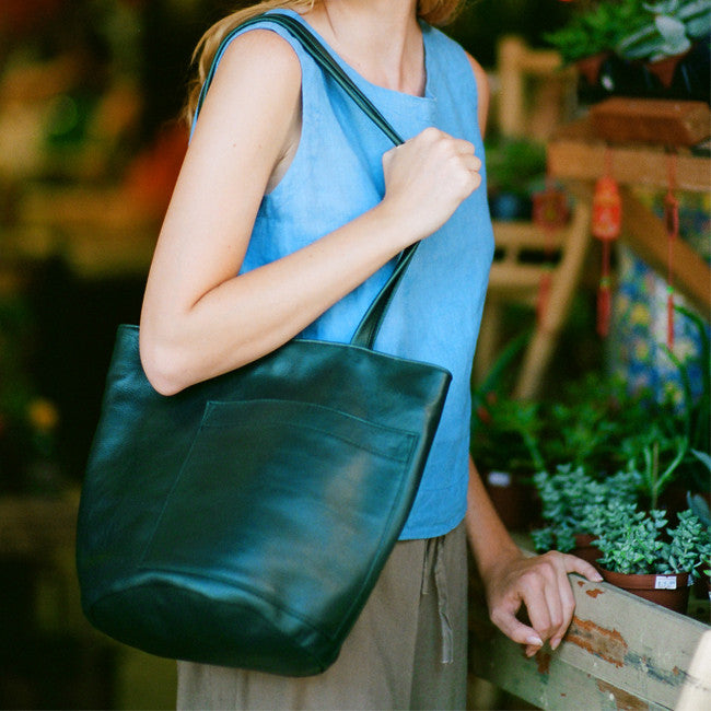 the erin templeton bucket bag in forest leather shown on a model wearing an erin templeton cerulean blue linen tank in the foreground, a flower market in the background