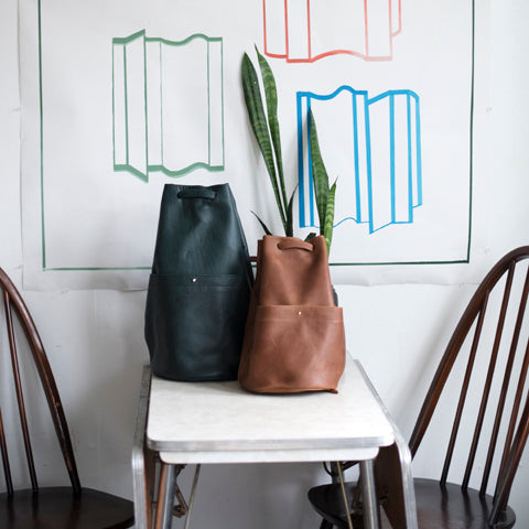 side by side large black and small chocolate gunny sack leather backpacks, both with a gathered strap closure and large front pocket with a stud closure, sitting on stop of a table in between two chairs and a painting hung on the wall behind