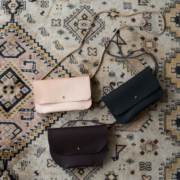 three small envelope style leather purses laying flat on an ornamental rug, shown in veg tan leather, chocolate leather, black leather, each with a stud closure. handmade by erin templeton in vancouver, canada