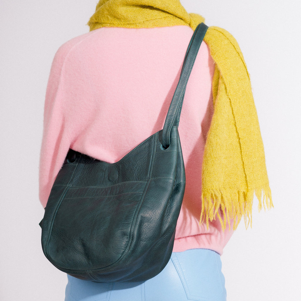 a leather tote with a large front pocket, double straps and a magnetic closure in forest leather, worn crossbody on a model in colourful clothing against a white backdrop