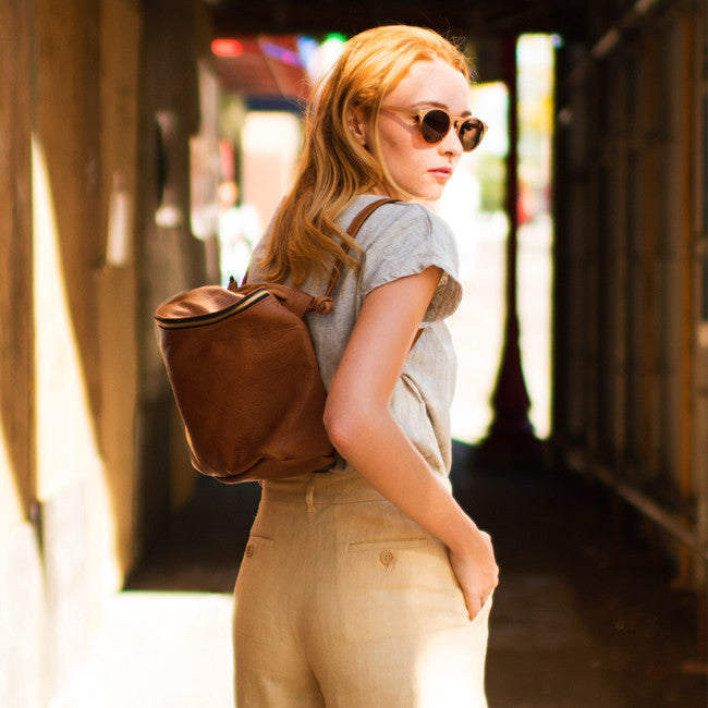 a small camera bag style leather backpack with a zip closure in caramel leather,  worn as a backpack on a model in a street setting