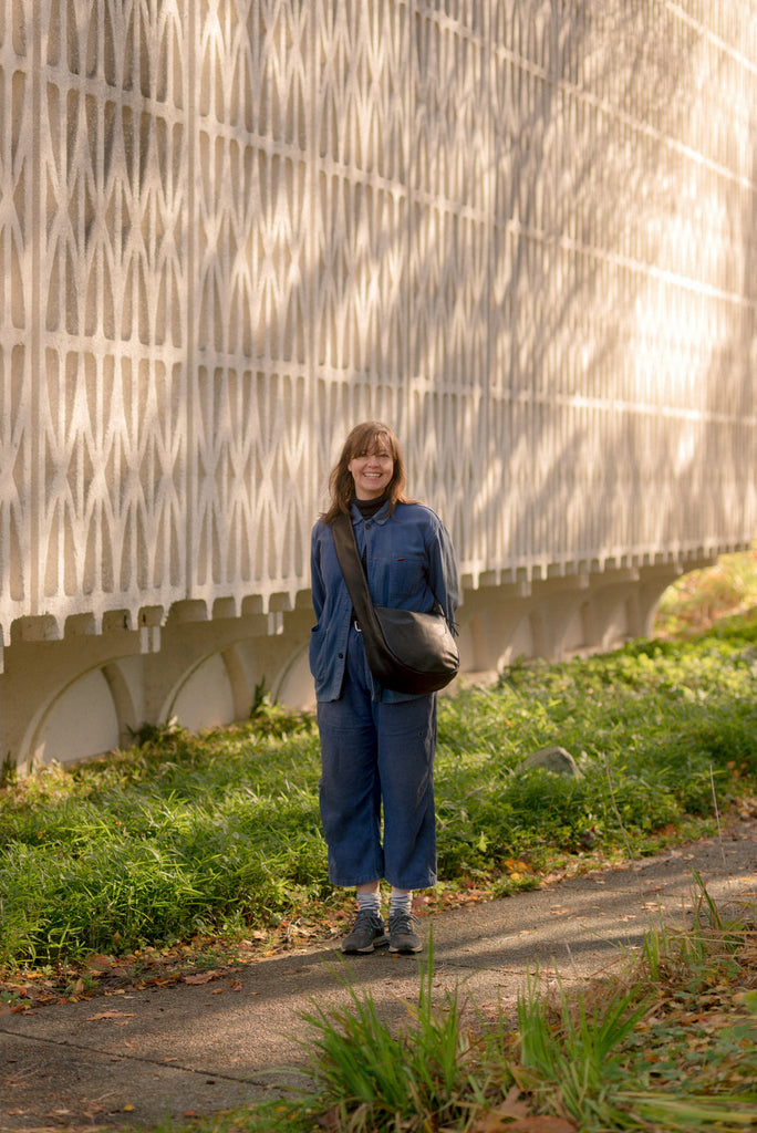 vancouver leather accessories designer Erin Templeton stands smiling in vintage denin and black leather daytripper bag she created. Erin stands next to sun dappled UBC campus building 