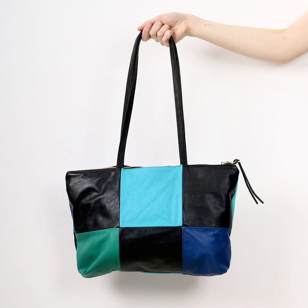 an outreached arm holding a patchwork style leather tote with 6 alternating panels of black and blue leather, a double zip closure and double straps, handmade by erin templeton in vancouver, canada