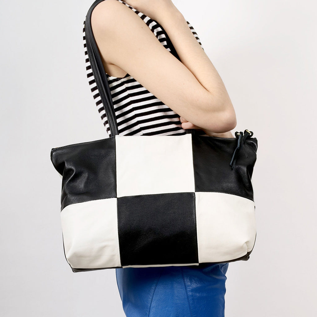 a patchwork style leather tote with 6 alternating panels of black and white leather, a double zip closure and double straps, worn on the shoulder of a model wearing a black and white striped top and blue leather pants in front of a white backdrop