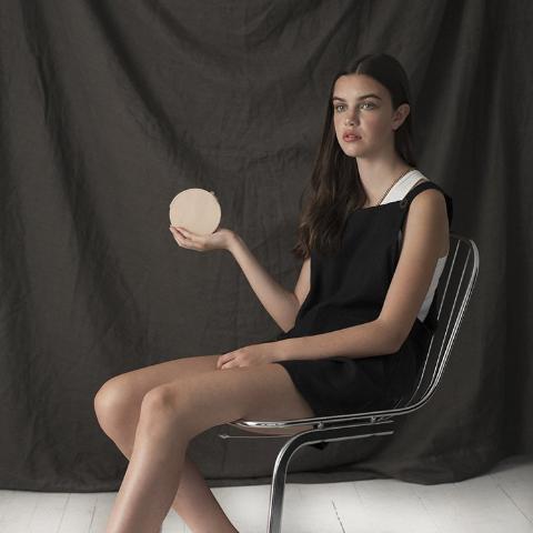 a model seated wearing erin templeton linen overalls and holding small leather coin purse in veg tan leather in front of a grey backdrop, handmade by erin templeton in vancouver, canada