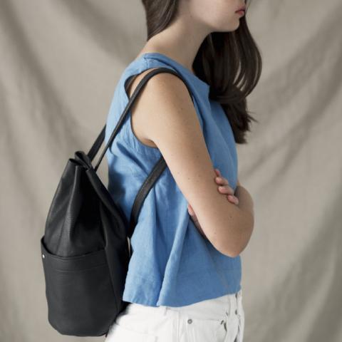 a small gunny sack leather backpack in black leather with a gathered strap closure and large front pocket with a stud closure,  shown from the side on a model wearing an erin templeton cerulean  blue linen tank in front of an off-white backdrop