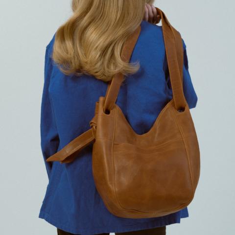 a large leather tote with a large front pocket, double straps and a magnetic closure in caramel leather, worn over the shoulder of a model in a blue jacket in front of a grey backdrop