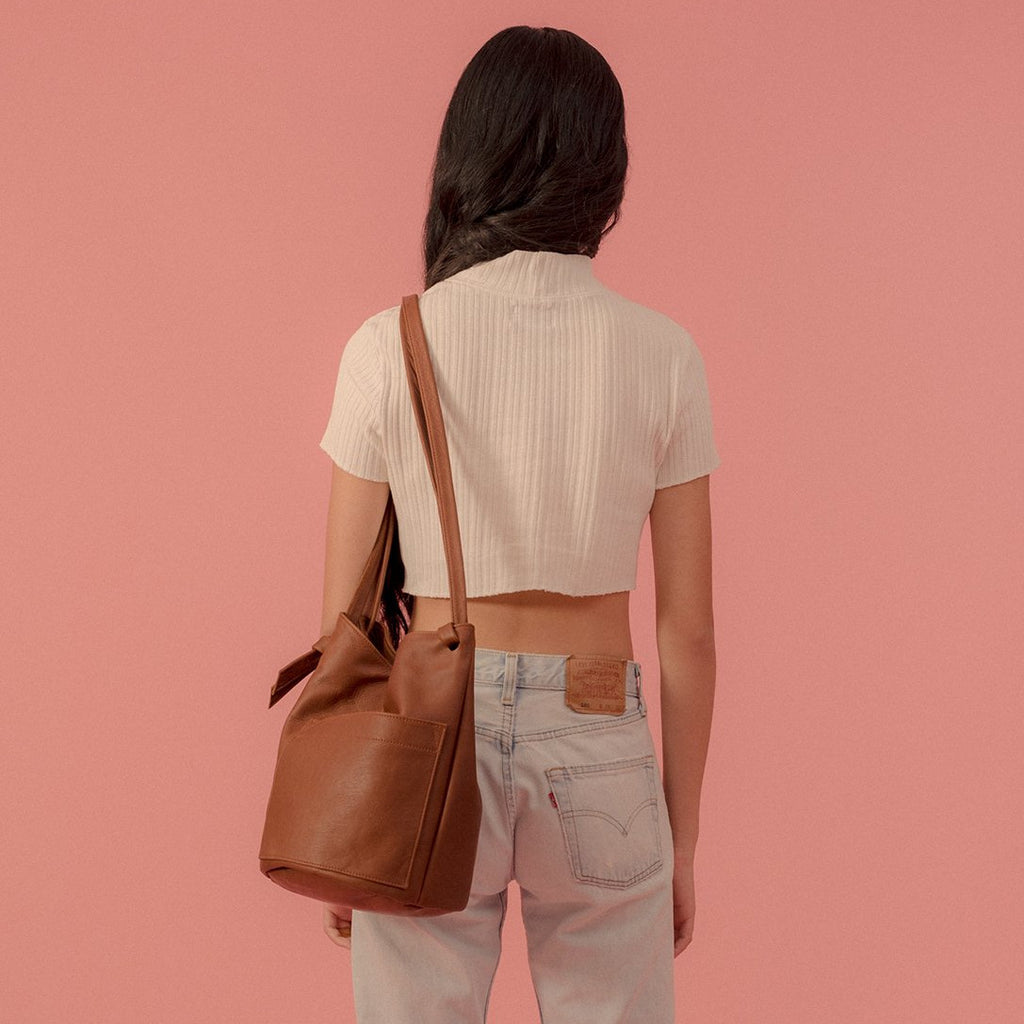 the erin templeton cross body bucket bag in caramel, shown on a model from behind on a pink background. the model is wearing a cream coloured ribbed tee and light wash levi's jeans
