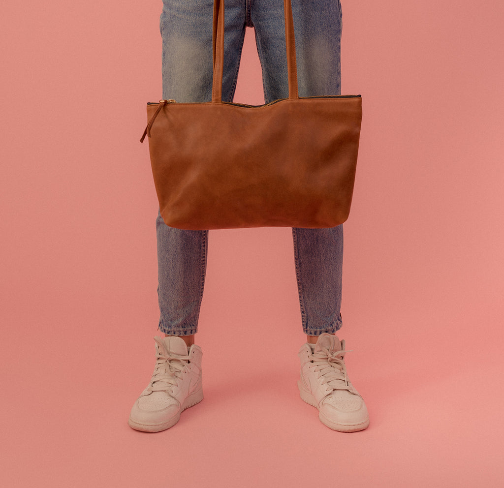 a large 'byob horizon' leather handbag with a zipper closure and secret pocket in caramel leather, shown in front of a models legs, wearing mid wash jeans and white sneakers on a  pink background