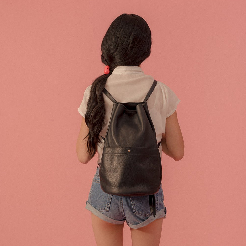 a large gunny sack leather backpack in black leather with a gathered strap closure and large front pocket with a stud closure, worn on a model in front of a pink background