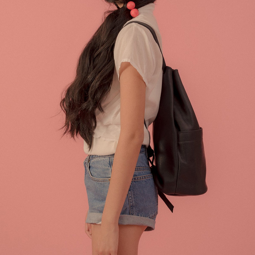 a large gunny sack leather backpack in black leather with a gathered strap closure and large front pocket with a stud closure, shown from the side on a model in front of a pink background