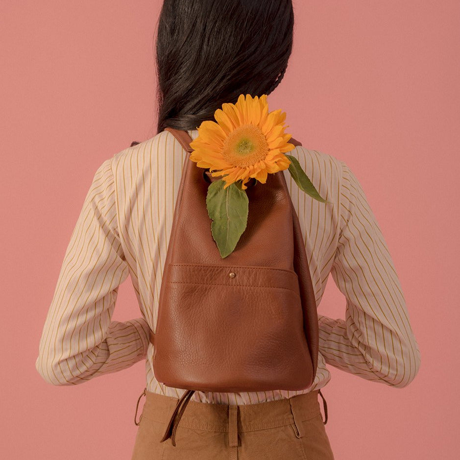 a small gunny sack leather backpack in caramel leather with a gathered strap closure and large front pocket with a stud closure and a sunflower pocking out of the top, worn on the back of a model in front of a pink back drop