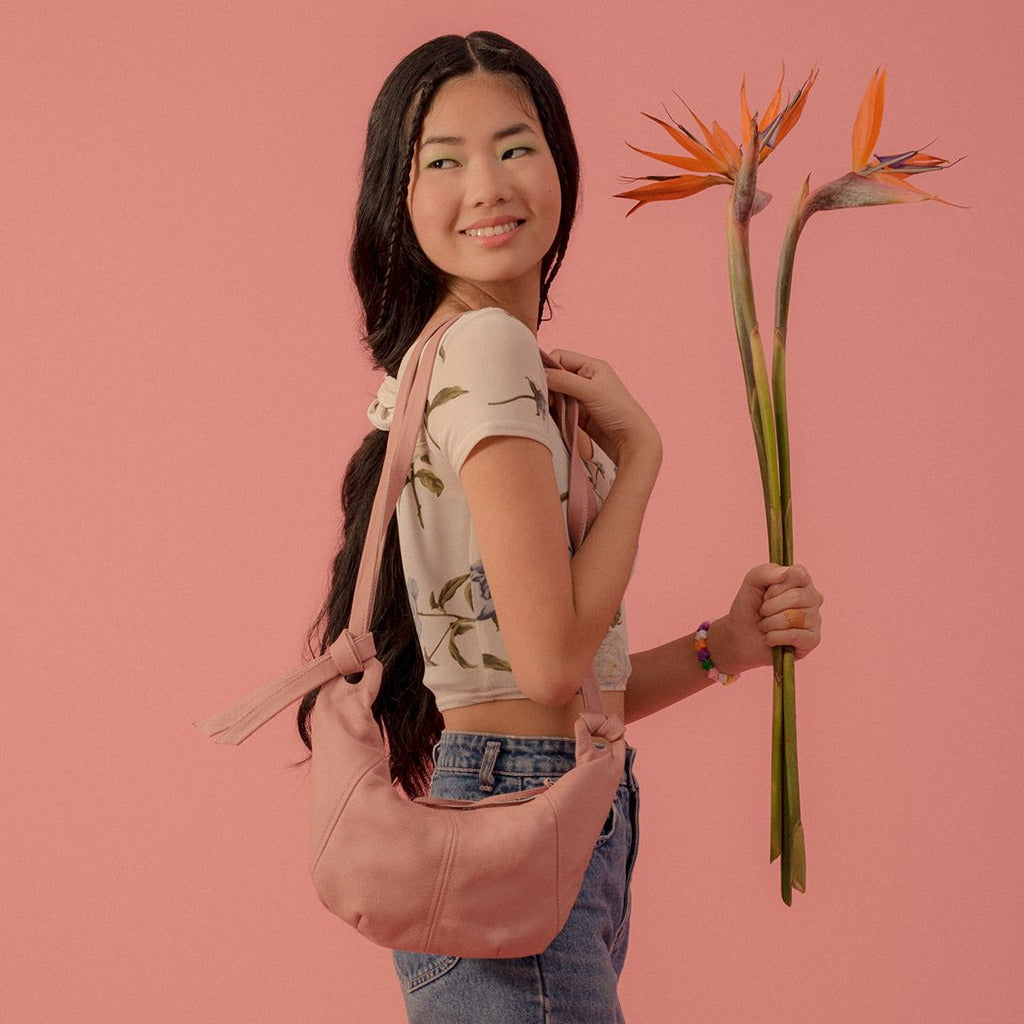a mini hobo leather purse in blush leather with a zip closure, worn over a models shoulder who is holding a bouquet of birds of paradise cranes and standing in front of a  pink backdrop