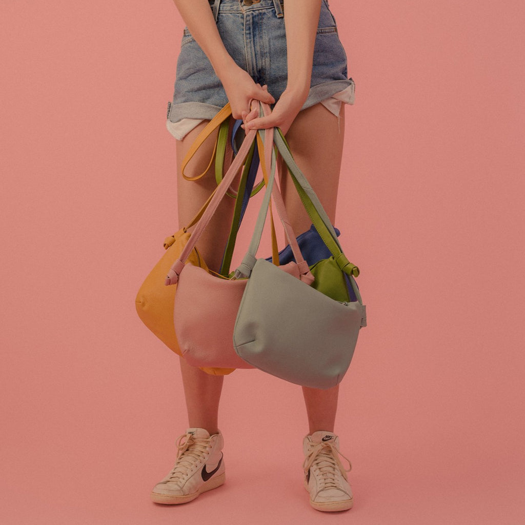 multiple tgif small leather purses shown in toothpaste, blush, mustard, kermit and blue leather, held by a model wearing denim shorts and nike sneakers against a pink background