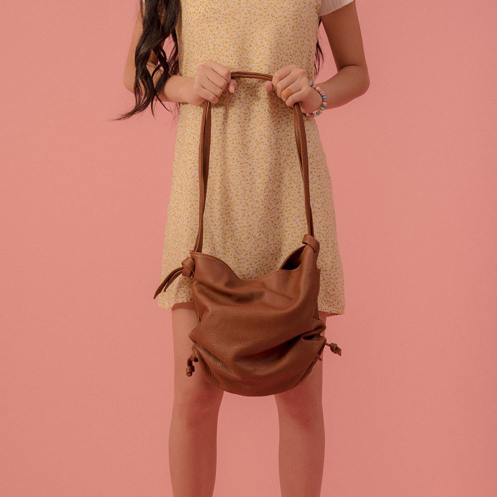 a large leather tote with adjustable ruched sides in caramel leather, held in front of a model in a yellow dress in front of a pink backdrop