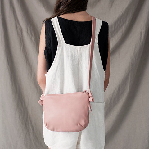 a small leather purse shown in blush leather, worn crossbody on a model in a white dress against a grey backdrop