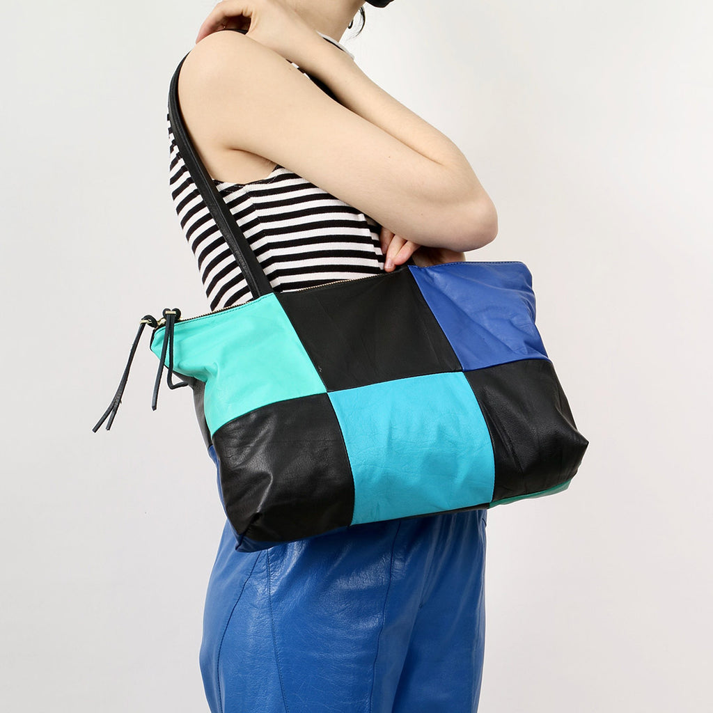 a patchwork style leather tote with 6 alternating panels of black and blue leather, a double zip closure and double straps, worn on the shoulder of a model wearing a black and white striped top and blue leather pants in front of a white backdrop