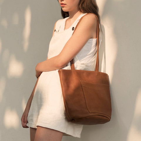 the erin templeton bucket bag in caramel leather, shown on a model wearing erin templeton linen orveralls leaning against an off-white wall