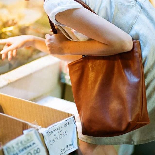 a 'byob square' leather tote with a magnetic closure and secret pocket in caramel leather, shown from the side on a women bending over  fresh pasta at a market