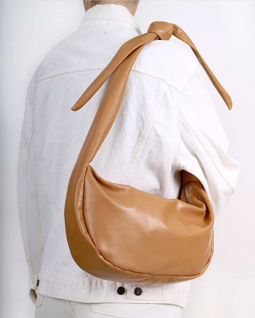 a large leather sling style handbag shown in camel leather with a knotted strap and double zip closure, worn on the shoulder of a model wearing a white denim jacket in front of a white backdrop