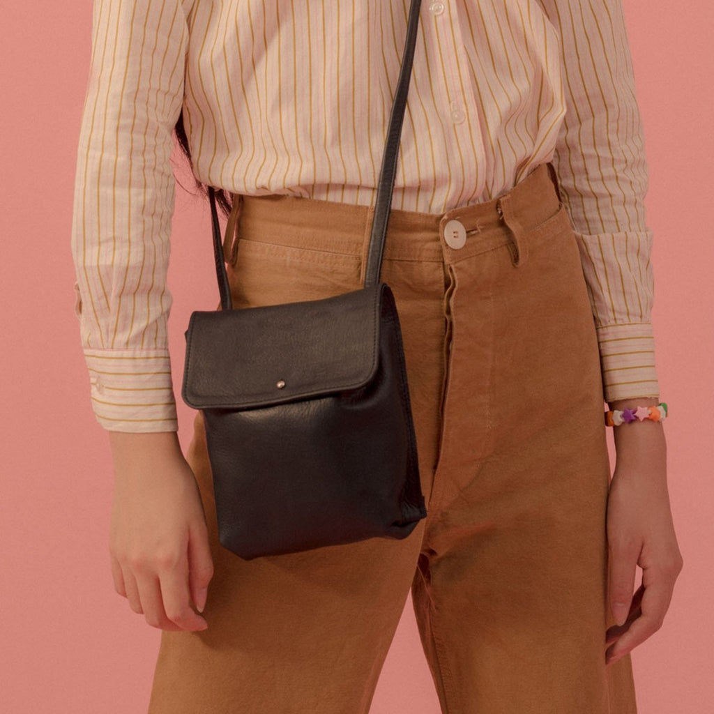 a small cross body leather purse with an envelope style flap and stud closure, shown in black leather on the hip of a model wearing a striped shirt and tan slacks in front of a  pink backdrop