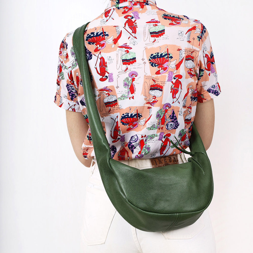 a small leather sling style handbag shown in sage leather with a knotted strap and double zip closure, worn cross body on a model wearing a printed shirt and white jeans in front of a white background