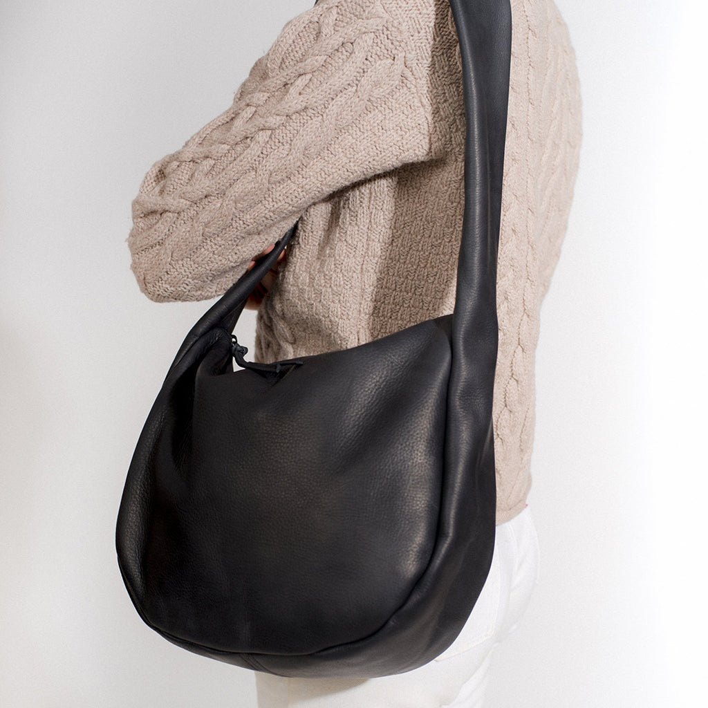 the day tripper leather satchel shown with a round body in black leather with a zip closure, shown on a model wearing an oatmeal cable knit sweater on a white background