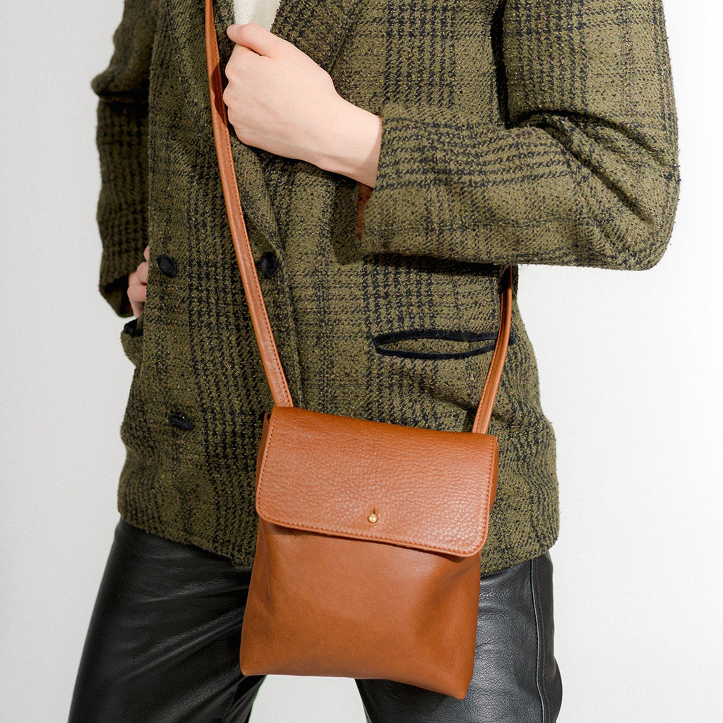 a small cross body leather purse with an envelope style flap and stud closure, shown in caramel leather on the hip of a model wearing a double breasted blazer and black leather pants in front of a white backdrop