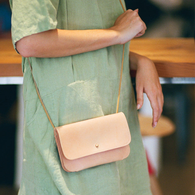 a small leather envelope style purse with a stud closure in veg tan leather, worn crossbody on a woman leaning against a table in a green dress