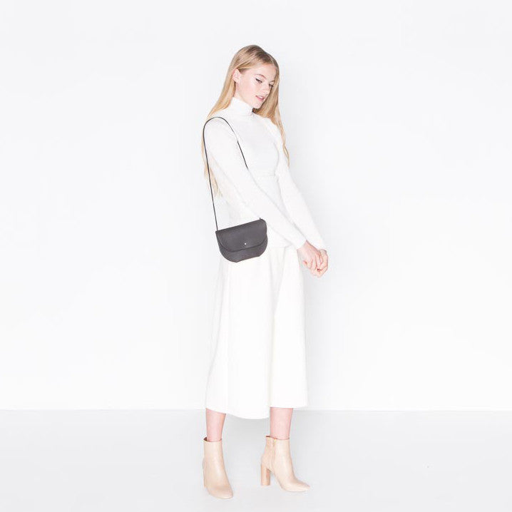 a small leather envelope style purse with a round bottom and stud closure in black leather, shown on the shoulder of a model wearing all white against a white backdrop