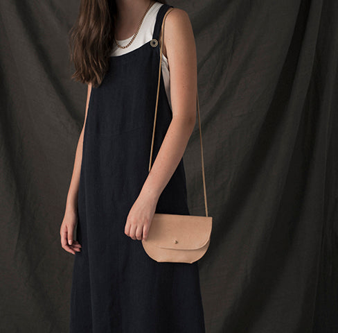 a small leather envelope style purse with a round bottom and stud closure in veg tan leather, shown on the shoulder of a model wearing a black dress against a grey backdrop