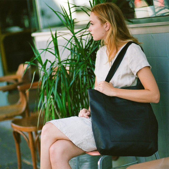 a large leather tote with a large centred patch-style pocket shown in black leather, worn on the shoulder of a model sitting on a chair outside a business next to a plant